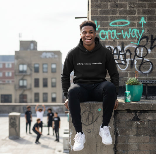 Man seated outdoors with graffiti wall, wearing a black cra-way math delulu hoodie styled with casual streetwear